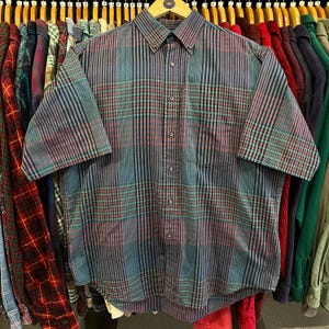 May include: A short-sleeved button-down shirt with a plaid pattern in shades of blue, red, and black. The shirt has a button-down collar and a single chest pocket. The shirt is hanging on a wooden hanger.