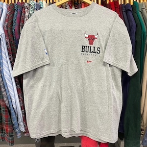 Pu&ograve; includere: Una maglietta grigia con il logo dei Chicago Bulls in rosso e nero. La parola "Bulls" &egrave; stampata sotto il logo, con "Basketball" sotto. &Egrave; visibile un piccolo logo Nike.