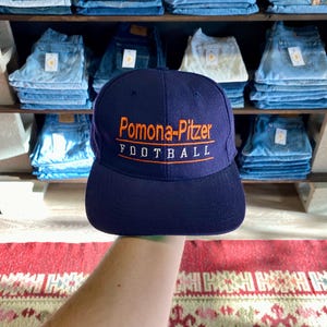 May include: A navy blue baseball cap with the text "Pomona-Pitzer FOOTBALL" embroidered in orange. The cap is held up in front of a shelf of folded jeans. The background includes a red and white patterned rug.