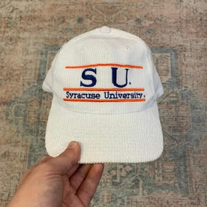 May include: White corduroy baseball cap with the letters "SU" embroidered in blue and orange, above "Syracuse University". The hat has a curved brim and a structured crown.