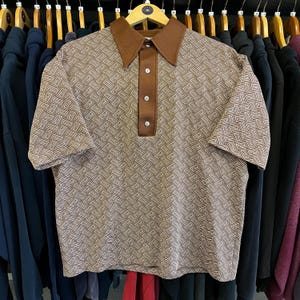 May include: A short-sleeved polo shirt with a geometric pattern in shades of brown and white. The shirt features a brown collar and a button-up placket. The shirt is hanging on a wooden hanger.