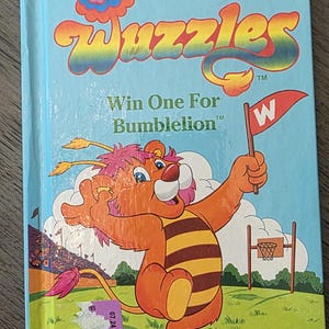 Op de afbeelding: Een vintage kinderboek getiteld "Wuzzles Win One For Bumblelion". De cover toont een cartoonachtig leeuwachtig personage dat een vlag met een "W" vasthoudt. Het boek maakt deel uit van de Collector Series.
