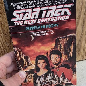Puede incluir: Un libro de bolsillo titulado "Star Trek: The Next Generation: Power Hungry" de Howard Weinstein. La portada presenta un paisaje con dos personas y el logotipo de Star Trek. El libro está sostenido por una persona.