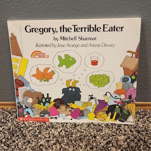May include: A children's book titled "Gregory, the Terrible Eater" by Mitchell Sharmat, illustrated by Jose Aruego and Ariane Dewey. The cover features a goat and thought bubbles with food items. The book is published by Scholastic.
