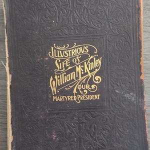 Puede incluir: Un libro negro con letras doradas en la portada. El título dice "Illustrious Life of William McKinley Our Martyred President".