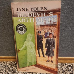 May include: Book cover for "The Devil's Arithmetic" by Jane Yolen. The cover features a green door with a ghostly figure of a young woman opening it. Behind the door, a group of people are standing in a line, and a guard in a uniform is watching them. The book has won the National Jewish Book Award.