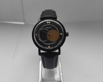 Puede incluir: Reloj Raketa negro con esfera redonda y correa de cuero negra. La esfera del reloj presenta un diseño negro y dorado con la palabra "RAKETA" impresa. El reloj se exhibe sobre un soporte transparente.