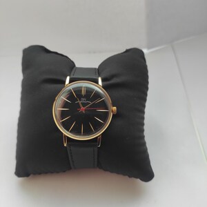 May include: A gold-toned wristwatch with a black face and gold hands, resting on a black pillow. The watch has a black leather band. The watch face has gold markers and a red second hand.