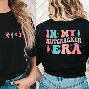 Puede incluir: Camiseta negra con un gráfico de bailarines en la parte delantera y el texto "IN MY NUTCRACKER ERA" en la parte posterior. El texto está en una fuente retro y presenta una silueta de cascanueces.