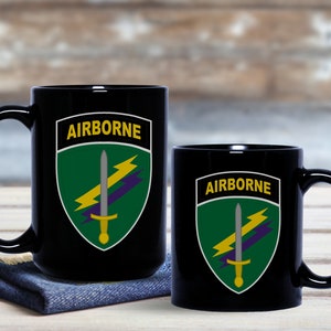 May include: Two black coffee mugs with a green, yellow, and purple military insignia. The insignia features a sword and lightning bolts. The text "AIRBORNE" is printed on each mug.