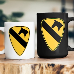May include: Two ceramic coffee mugs with a black and yellow military insignia. The insignia features a horse's head and a black diagonal stripe on a yellow background.