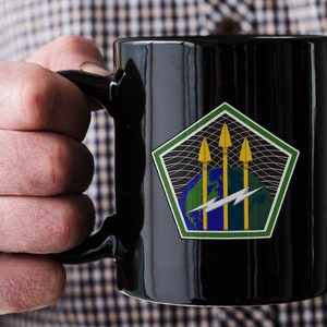 May include: A black coffee mug with a green, blue, and yellow graphic of a pentagon with three arrows and a lightning bolt. The graphic is on a white background.