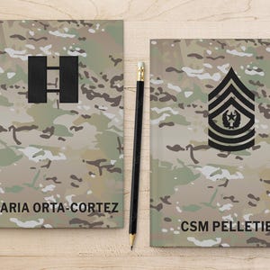 May include: Two camouflage notebooks with black text and insignia. The notebook on the left has the text "MARIA ORTA-CORTEZ" and a black insignia of two horizontal bars. The notebook on the right has the text "CSM PELLETIER" and a black insignia of a star with wings.