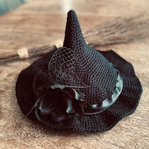 May include: A black witch hat with a wide brim, a pointed crown, and a black ribbon. The hat is decorated with a black flower and a black mesh veil.