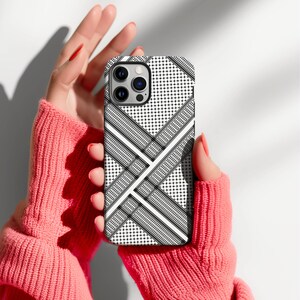 May include: A black and white patterned phone case with a geometric design. The case features a repeating pattern of small dots, diagonal lines, and wavy lines. The phone case is held by a person wearing a pink sweater.