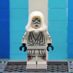 Pode incluir: Minifigura Lego de um personagem com um traje branco com capuz e detalhes pretos. A figura tem um rosto detalhado com olhos verdes e uma máscara preta. A figura está em frente a um fundo azul e branco.