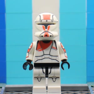 May include: A white and red Lego minifigure in a battle suit. The figure has a white helmet with red accents, a red scarf, and black hands. The figure is standing in front of a blue and white striped background.