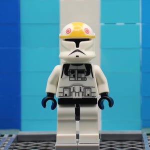 May include: A white Lego minifigure of a Star Wars clone trooper with a yellow helmet and black markings. The figure has blue hands and is standing in front of a blue and white background.