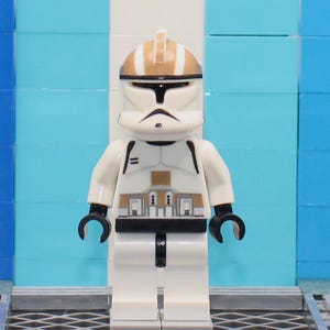 May include: A white and tan Lego minifigure of a clone trooper, with black accents and a helmet. The figure stands in front of a blue and white background. The figure has a black visor and a black mouth.