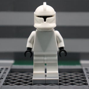 May include: A white Lego minifigure of a clone trooper, standing upright. The figure has a white helmet with a black visor, white torso, white legs, and black hands. The background is a blurred gray and green.