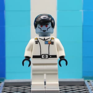 May include: A Lego minifigure of a character with blue skin, red eyes, and black hair. The figure wears a white uniform with black accents, including a belt and gloves. A small badge with colorful details is on the chest.