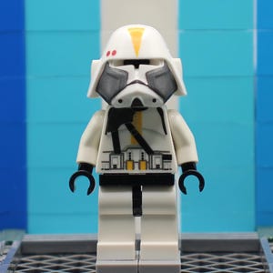 May include: A white Lego minifigure of a Star Wars clone trooper with a helmet featuring yellow and red accents. The figure has black details on the chest and arms, and black hands. The background is a mix of blue and white.