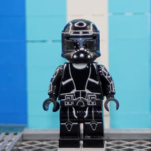Könnte beinhalten: Eine schwarze Spielzeug-Minifigur aus Kunststoff mit Helm und Rüstung, die mit feinen weißen Linien verziert ist. Die Figur hat ein dunkles Visier, ein segmentiertes Rumpfdesign und bewegliche Gliedmaßen auf einer kleinen grauen Grundplatte.