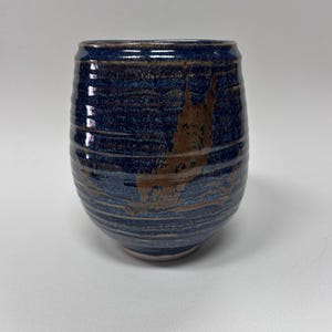 Puede incluir: Un vaso de cerámica hecho a mano con un esmalte azul oscuro y marrón. La taza tiene una forma redondeada y una superficie texturizada con líneas horizontales. El interior es de un tono azul más claro.
