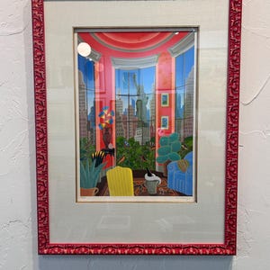 May include: Framed artwork featuring a vibrant interior scene with a view of the New York City skyline, including the Statue of Liberty. The artwork is framed in a red ornate frame. The interior includes a yellow chair, blue sofa, and potted plants.