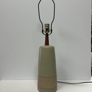 May include: A mid-century modern table lamp with a ceramic base in shades of green and tan. The lamp features a wooden neck and finial, a metal lamp shade holder, and a black power cord. The lamp is designed for a standard light bulb.
