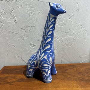 Może przedstawiać: Niebieska ceramiczna figurka żyrafy z białymi wzorami w kształcie liści. Żyrafa ma długą szyję i stoi na czterech nogach. Figurka stoi na drewnianej powierzchni.