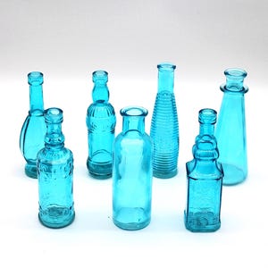 Könnte beinhalten: Sieben kleine, blaue Glasflaschen in verschiedenen Formen und Größen. Die Flaschen sind auf einer weißen Oberfläche angeordnet.