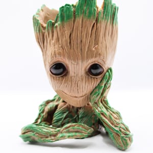 Puede incluir: Un macetero de cerámica verde y marrón con forma del personaje Groot de la película Guardianes de la Galaxia. El macetero está sobre una superficie blanca y tiene un pequeño agujero en la parte superior para el drenaje.