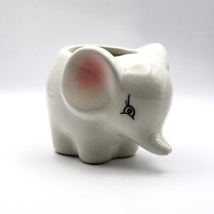 Könnte beinhalten: Weißer Keramik-Elefantentopf mit rosa Ohr und schwarzem Augendetail.