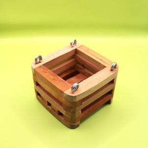 May include: A small, square wooden planter with a natural finish. The planter has a slatted design and metal hanging loops. The wood tones range from light to medium brown. The planter is designed for small plants.