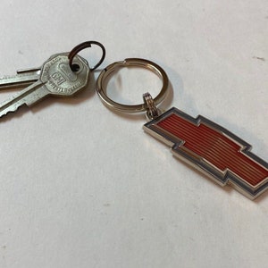 May include: A silver key ring featuring a red and silver Chevrolet logo, accompanied by two keys. The keys are silver and bear the GM logo. The key ring is circular and crafted from silver metal.