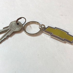 Puede incluir: Un llavero plateado con un dije de logotipo de Chevrolet amarillo y dos llaves plateadas. Las llaves tienen el texto "General Motors" y "GM" en ellas.