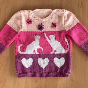 May include: A pink, purple, and white striped knitted sweater with a design of two cats and butterflies. The sweater has white hearts on the bottom.