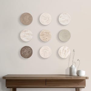Puede incluir: Nueve piezas decorativas redondas para la pared en tonos beige, crema y marrón se exhiben en una pared clara. Debajo hay una consola de madera con dos cajones. Dos pequeños jarrones blancos y un cuenco blanco pequeño están sobre la mesa.
