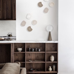 Könnte beinhalten: Ein modernes Wohnzimmer mit einem Schrank aus dunklem Holz, einer weißen Wand mit einer Sammlung von Keramikwanddekorationen und einer braunen Lampe auf einem Tisch.
