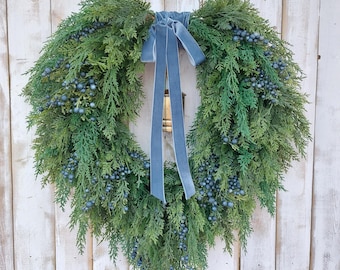 May include: A lush, green wreath adorned with clusters of blue berries and a soft, gray-blue velvet ribbon. The wreath is a full, circular design, perfect for seasonal decor. The background is a white, weathered wooden door.