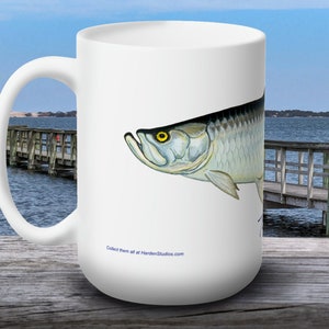 May include: White ceramic coffee mug with a colorful illustration of a tarpon fish. The text "Collect them all at HardenStudios.com" is printed on the bottom of the mug.
