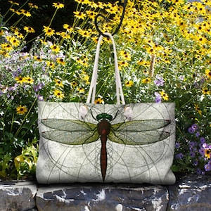 Puede incluir: Una bolsa de lona blanca con un diseño de libélula. La libélula es verde, roja y marrón con detalles intrincados. La bolsa tiene asas de algodón blanco.