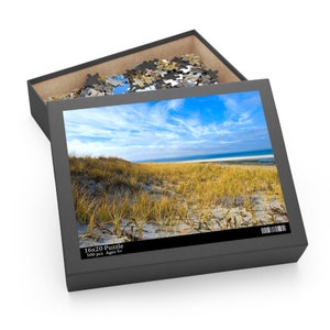 May include: A 500 piece jigsaw puzzle in a black box with a picture of a beach scene with sand dunes and a blue sky. The box says "16x20 Puzzle 500 pcs Ages 9+"