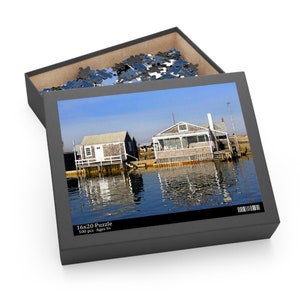 May include: A 500 piece jigsaw puzzle featuring a photograph of two wooden houses on stilts over a body of water. The houses are painted white and have brown roofs. The water is a light blue color and reflects the sky and the houses. The puzzle box is gray and has the text "16x20 Puzzle 500 pcs. Ages 9+" printed on it.