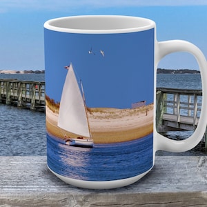 Peut inclure: Mug en céramique blanche avec un fond bleu représentant un voilier avec une voile blanche sur une étendue d'eau. L'eau est bleue et il y a une jetée en bois en arrière-plan.