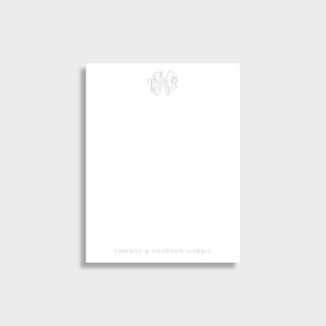 May include: A white notepad featuring a gold monogram at the top and the names "Thomas & Shannon Norris" printed at the bottom in a light brown colour. The monogram is an elegant, intertwined design.