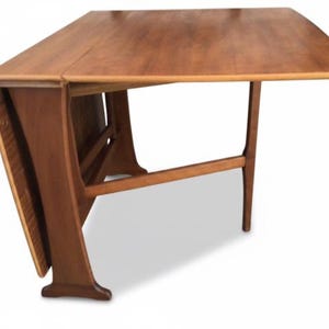 May include: A brown wooden drop-leaf table with a rectangular top and a hinged side panel that folds downwards. The table features a sturdy frame with angled legs and a horizontal support bar connecting the legs to the base of the tabletop.