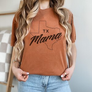 May include: A burnt orange t-shirt featuring the outline of the state of Texas with the words "TX Mama" in a stylish script. The shirt is made of a soft material and is worn with blue jeans.