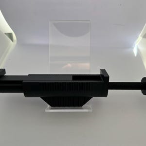 Może przedstawiać: Czarny, regulowany zacisk z żebrowanym uchwytem i gwintowanym prętem. Zacisk jest przeznaczony do utrzymywania przedmiotów na miejscu. Wykonany jest z czarnego tworzywa sztucznego i stoi na przezroczystym, akrylowym stojaku.